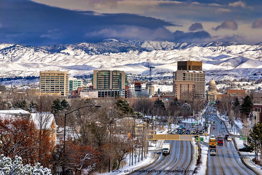 Photo of the city of Boise, ID during winter. In the right foreground is a street leading to the capitol building, with traffic including a school bus. In the middle of the image are Boise's buildings and in the background are snowy mountains.